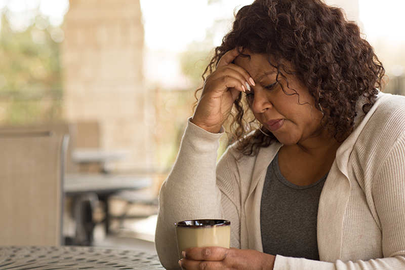 A mature woman looks stressed and worried as she holds her head in her hands and a cup of coffee