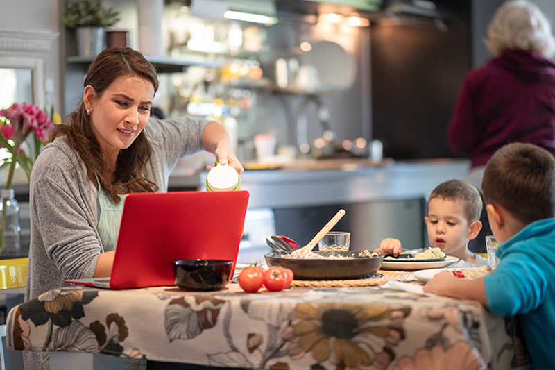 A woman works on her laptop as her two young children eat dinner and her elderly mother is in the background