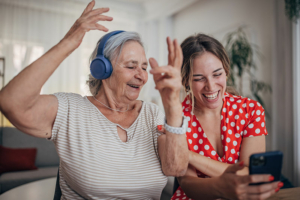 A caregiver utilizing music as one of a number of new ideas for dementia care listens to headphones with a happy older woman.