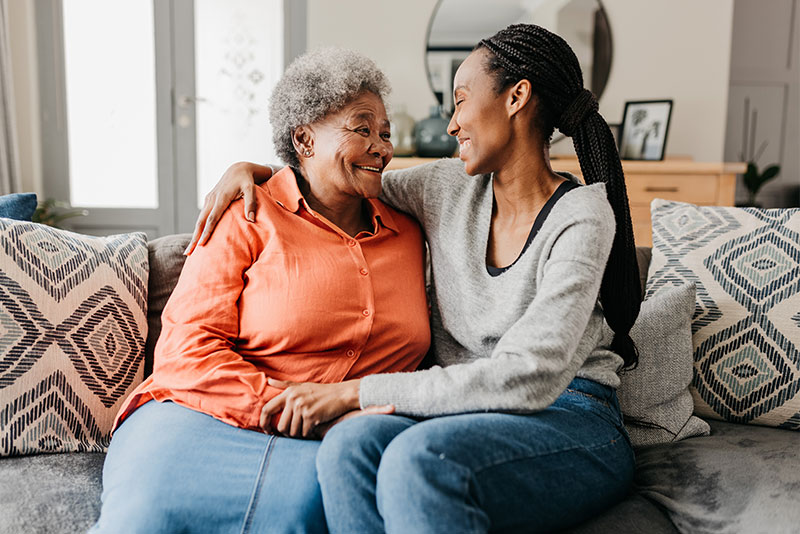 A woman who utilizes tips on how to help parents maintain independence wraps an arm around her older mother on the sofa as they both smile at each other.
