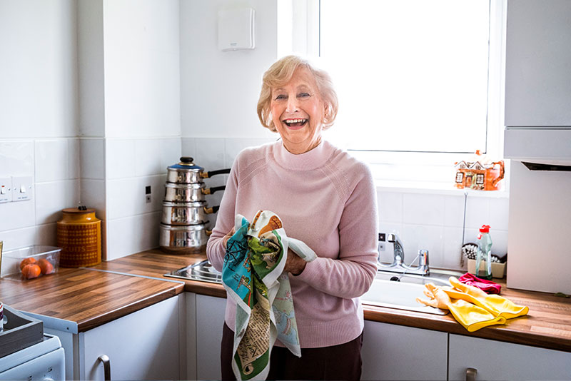An older woman smiles as she dries a dish in the kitchen, thanks to a caregiver who knew how to boost self-confidence for someone with dementia.