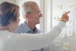 A woman utilizes one of many effective dementia care tips by posting reminder notes on her refrigerator, pointing them out to an older man.