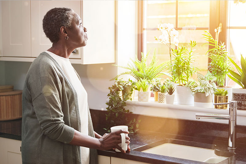 A woman who is feeling overwhelmed as a caregiver gazes out the kitchen window while holding a cup of coffee.