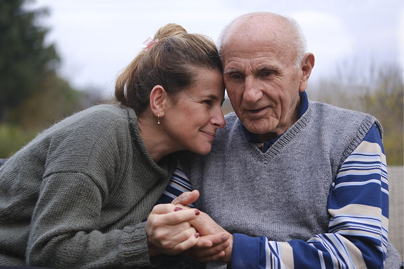 An older man struggling with Parkinson’s symptoms holds the hand of his caregiver as she gives him a hug.