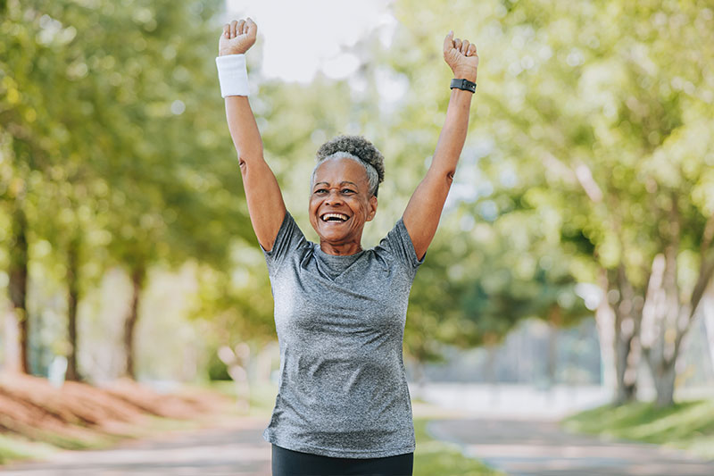 A woman who knows the importance of goal setting for seniors raises her arms in triumph as she completes a jog in the park.