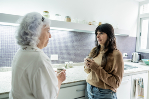 A woman resolving conflicts with an aging parent shares a warm laugh and coffee at the kitchen table with her parent.